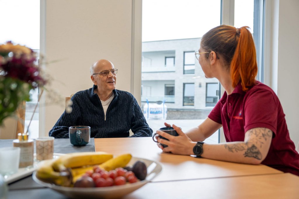 Man sieht einen freundlichen Kunden der Senioren-WG Springbachhöfe Paderborn im Gespräch mit einer Mitarbeiterin bei einer Tasse Kaffee.